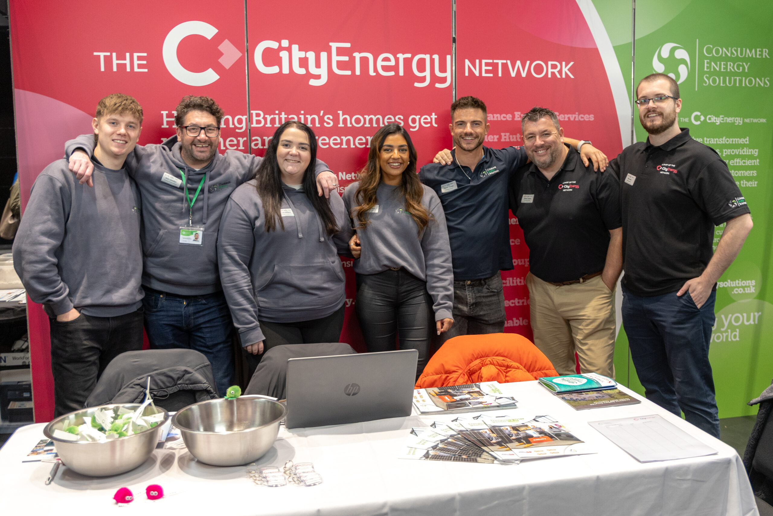 A group of colleagues stood in front of red branding with the words 'The City Energy Company' on. They are all wearing CES branded clothing and are a mix of gender, race and age.